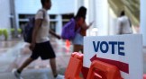 Two students in backpacks walk past a sign that says "Vote" at a polling location.