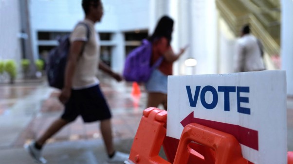 Two students in backpacks walk past a sign that says "Vote" at a polling location.