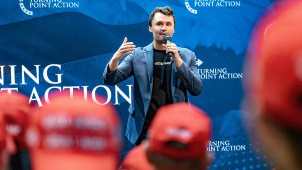 Charlie Kirk speaks with a microphone while people with red MAGA hats watch from the crowd.
