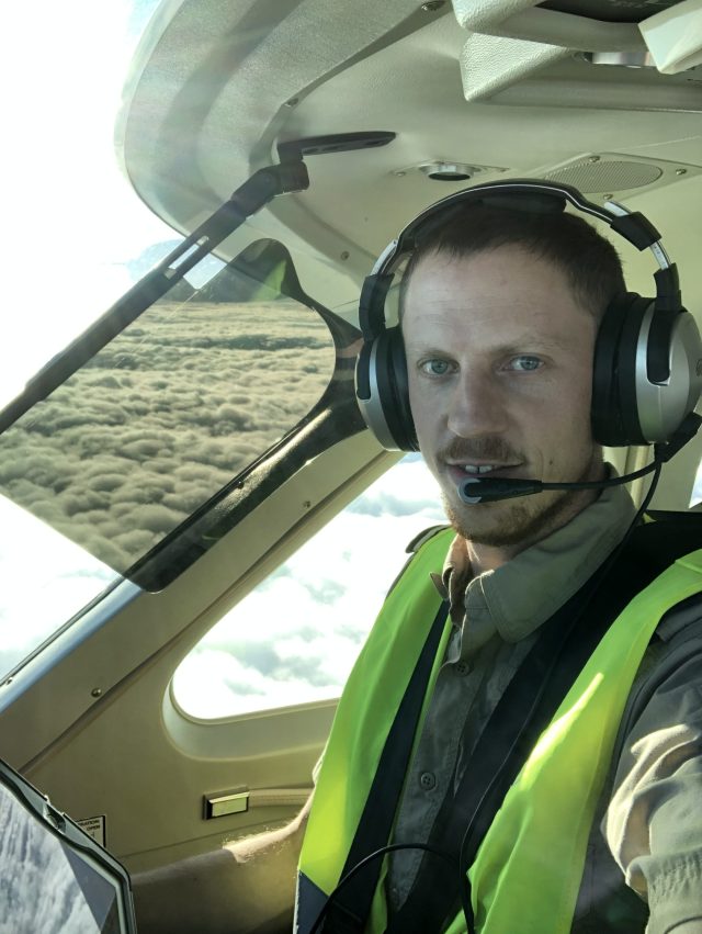 Ryan Koher wearing a yellow vest and headphones flying in a cockpit with clouds out the window.