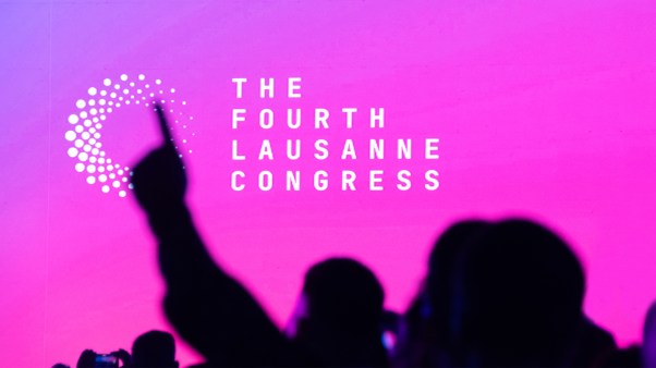 A hand reaching up in worship among a shadowed crowd in front of a pink screen with Lausanne on it